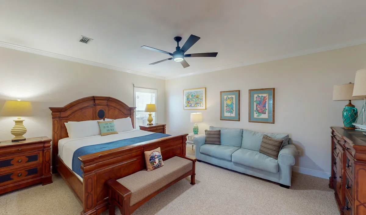 A spacious bedroom with a king-size bed, plush bedding, a cozy sofa positioned near the window, a nightstand with a lamp, and artwork on the wall. The room is filled with natural light, creating a serene and comfortable atmosphere ideal for relaxation.