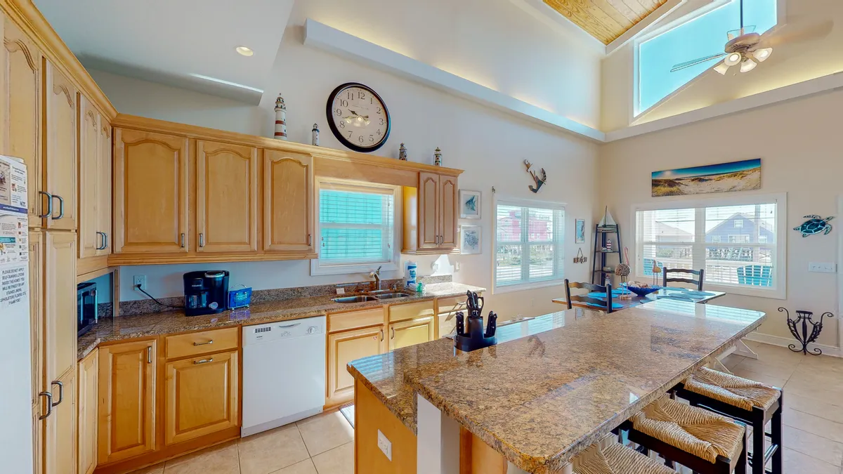A bright kitchen scene with a granite countertop, kitchen island seating, large windows, and colorful wall art. The layout is inviting, perfect for cooking, socializing, and preparing regional seafood or barbecue dishes.