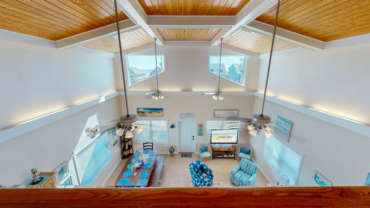A bright, welcoming open-plan living and dining area with vaulted ceilings, blue accent furniture, large windows flooding the space with natural light, and a visible entertainment area. The space feels airy and social, evoking a sense of togetherness and lively group gatherings.