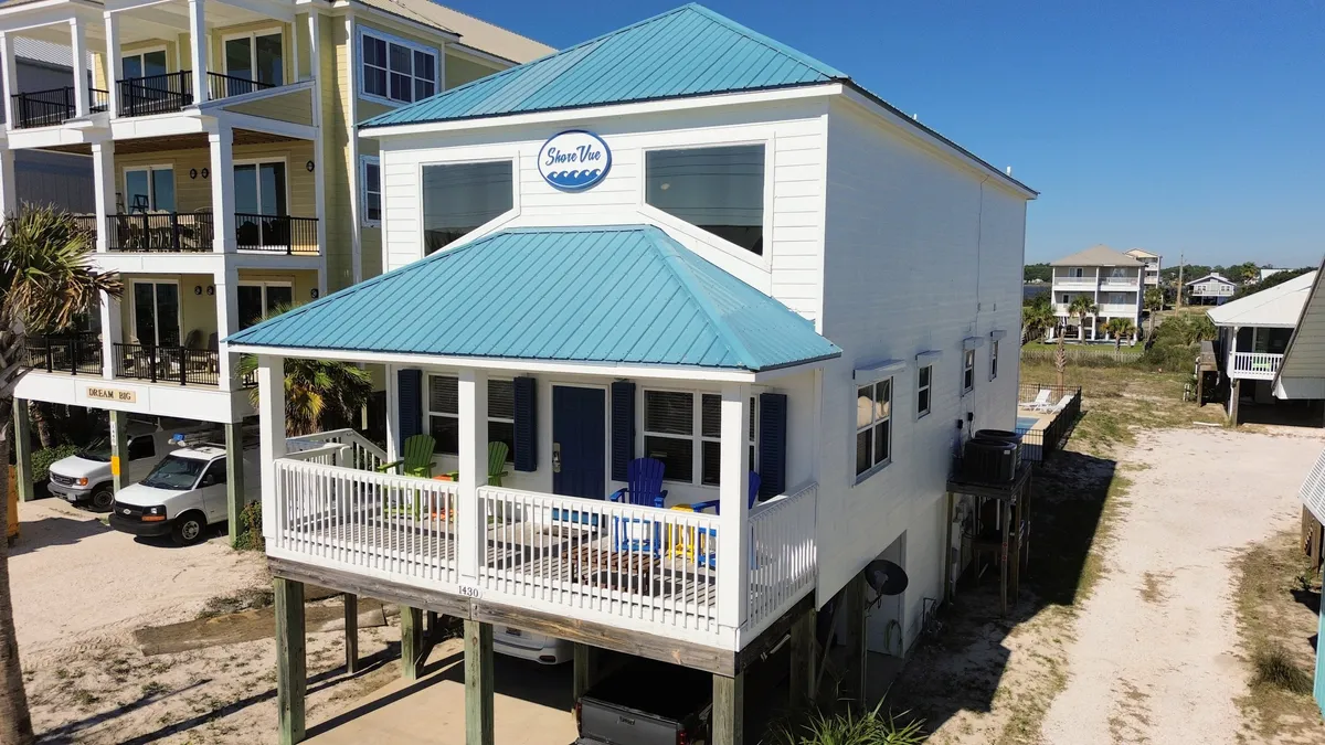 This image showcases the exterior of the home, featuring a blue roof, white siding, a welcoming porch with chairs, and the 'Slow Vee' house sign. The porch invites guests to relax and enjoy ocean views, echoing the feeling of returning to a private retreat after a day of exploring.