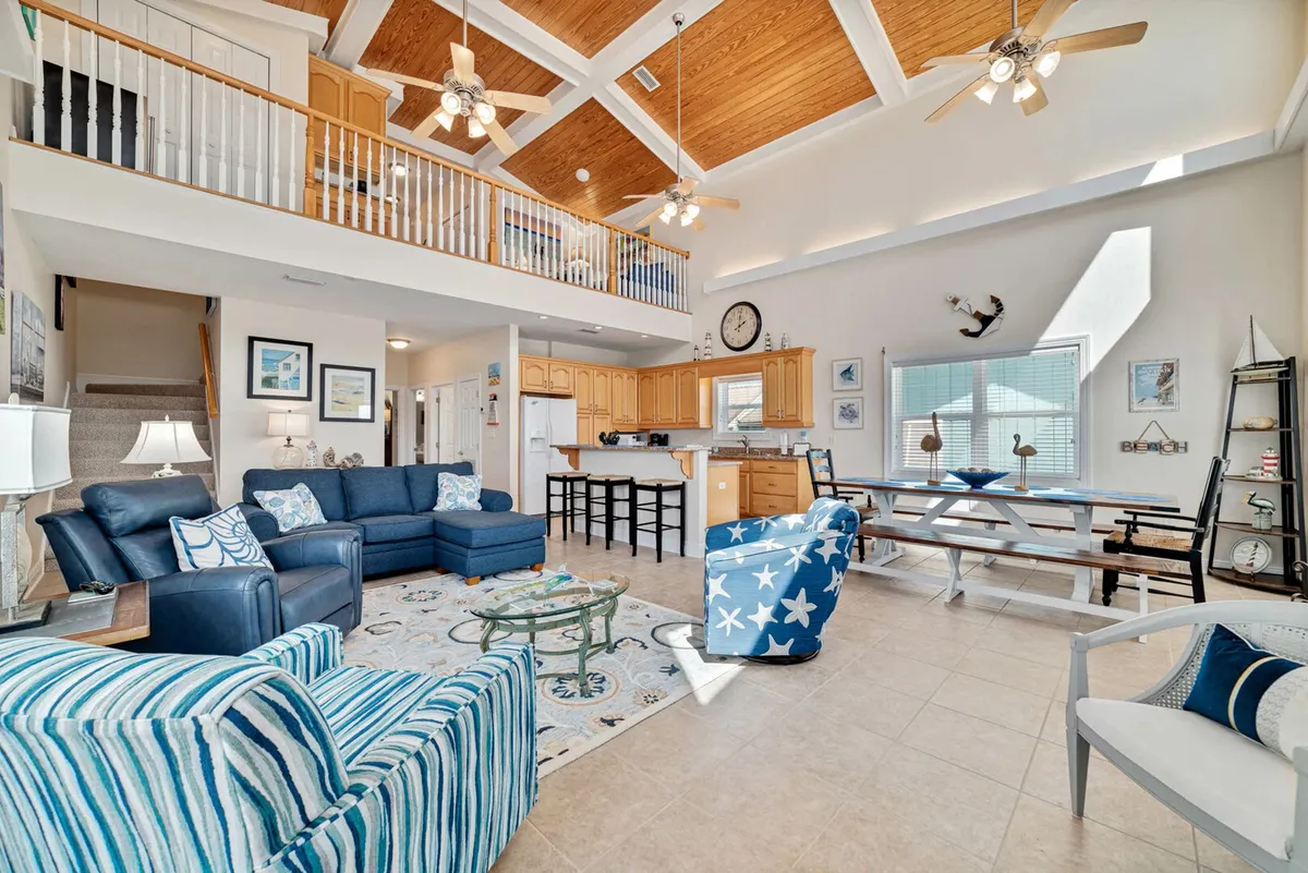 This image captures a spacious, open-plan living area featuring a large sectional sofa, striped accent chairs, a dining table, kitchen bar, and large windows that flood the space with natural light. The layout highlights easy flow between the living, dining, and kitchen areas, with comfortable furnishings and ample seating for group relaxation and entertaining.