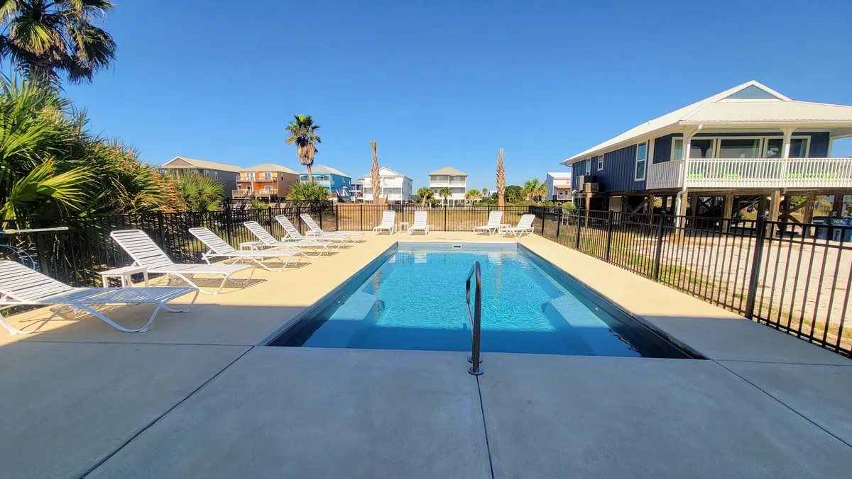 A refreshing swimming pool sits amidst lush palm trees, flanked by lounge chairs that invite sunbathing and relaxation. The houses in the background evoke a community setting and suggest the pool as a social hub for both families and friends.