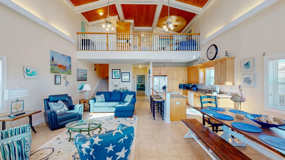 The hero image showcases a bright, airy open-concept living space with vaulted wooden ceilings, a stylish loft balcony, plush blue and white coastal furnishings, and a modern kitchen with a spacious breakfast bar. Sunlight floods the room through large windows, highlighting inviting seating areas perfect for family gatherings, meals, and relaxation, all accented by nautical décor that captures the Gulf Shores vibe.