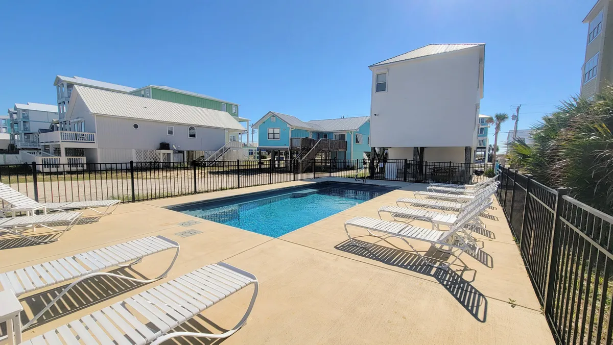A clear, inviting view of the rectangular private swimming pool with white lounge chairs arranged around its perimeter. The pool is securely fenced, and the background features modern coastal homes elevated on stilts, reinforcing the vacation vibe. The area is spacious, perfect for family gatherings and group play.