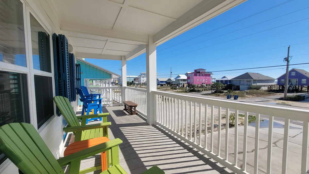 An exterior shot featuring colorful Adirondack chairs on the porch, with white railings casting shadows and a view of vibrantly painted beach houses across the street. The porch is bathed in sunlight, ideal for relaxing and soaking in the Gulf Shores atmosphere.