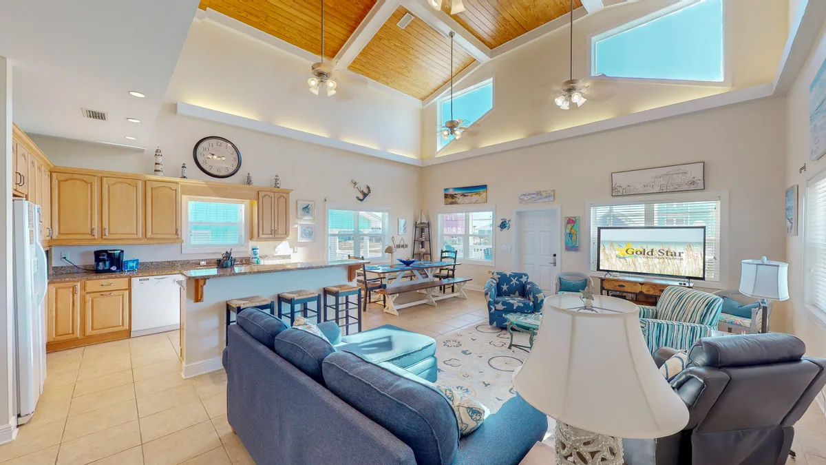 A sweeping perspective of the main social spaces, featuring a large kitchen with a breakfast bar, a spacious dining table with benches, and a comfortable seating area with couches and armchairs. The high vaulted ceiling and natural light create an uplifting environment ideal for family gatherings and entertaining.