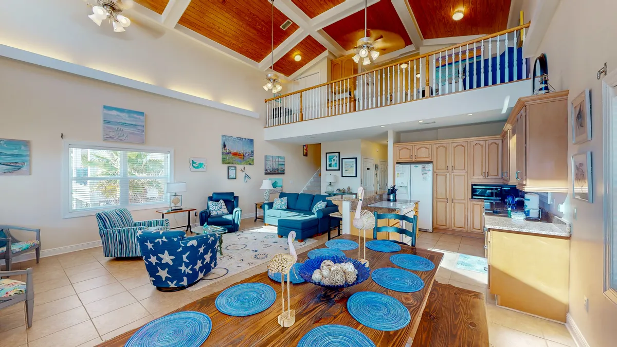 This image presents a spacious open-concept area that includes a large wooden dining table set with blue placemats and coastal decor, an adjacent open living space with blue and white nautical-themed seating, and a well-equipped kitchen with light wood cabinetry. The high vaulted ceiling with wood paneling and ceiling fans, as well as abundant natural light, reinforce the inviting, coastal atmosphere.