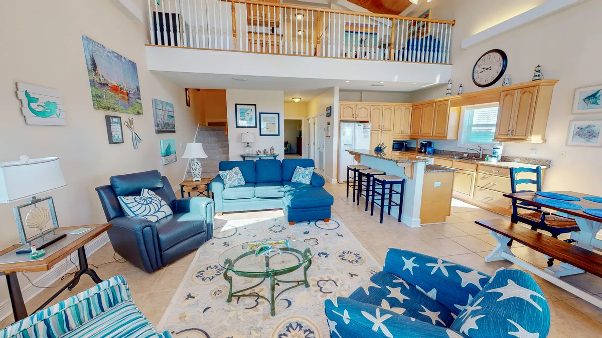 A cozy living room featuring a blue sectional sofa, glass coffee table, and coastal wall art. The kitchen with a breakfast bar and stools sits just behind the living area, making it easy to imagine relaxing, watching TV, or chatting over coffee. The overall palette and layout reinforce a sense of relaxation and togetherness.