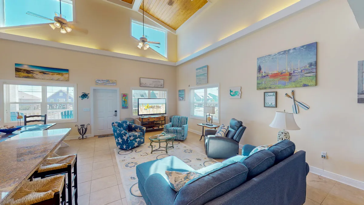 An interior living area with a large vaulted wooden ceiling, blue and patterned seating, nautical-themed decor, and expansive windows letting in natural light. The space is arranged for group gatherings, with a cozy atmosphere ideal for board games, movie nights, or socializing.