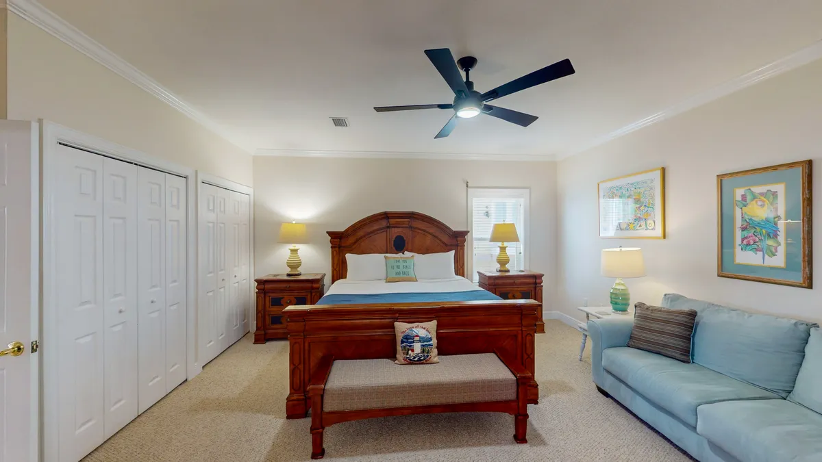 A serene king suite featuring a large wooden king-size bed with an ornate headboard, matching wooden nightstands with lamps, a light blue sofa with a striped pillow, and decorative framed coastal artwork. Soft lighting and plush bedding create a peaceful, inviting atmosphere ideal for rest.
