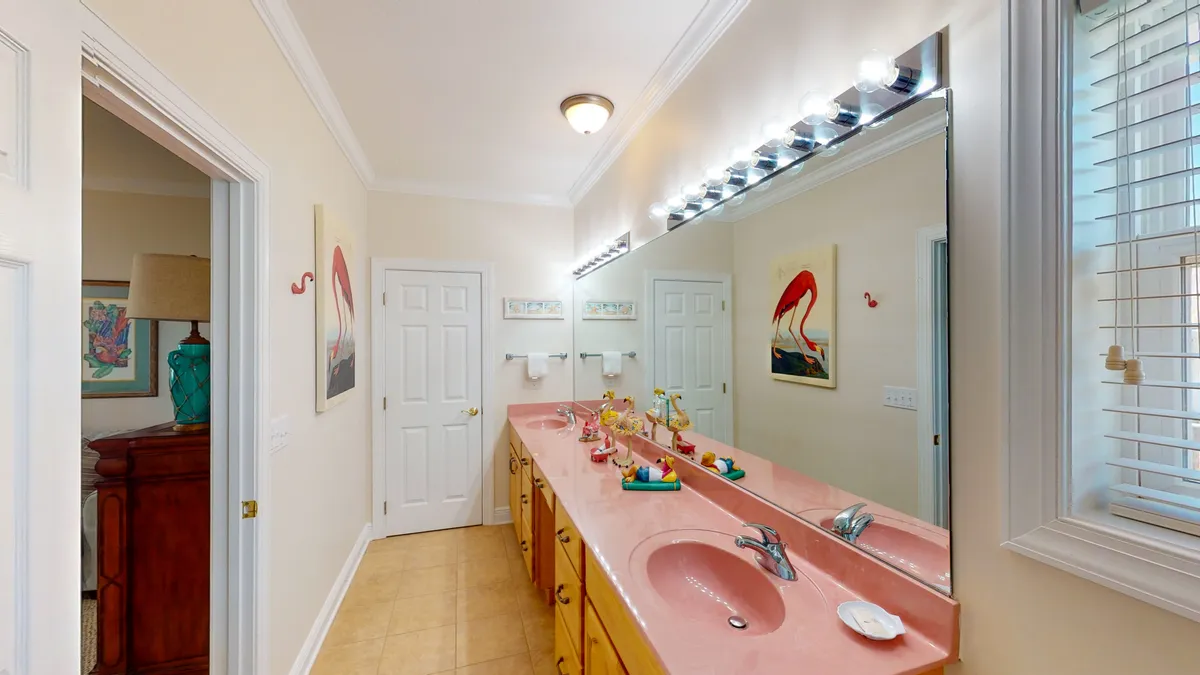 A bright bathroom with a long pink vanity countertop featuring dual sinks, a large wall mirror, flamingo artwork, and cheerful overhead lighting. The space appears meticulously clean, spacious, and welcoming for morning or evening routines.