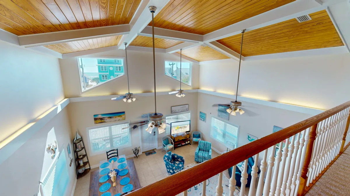 A bright, open living area with a vaulted wood-paneled ceiling, large windows that flood the room with natural light, a dining table set with blue accents, and a cozy seating area with nautical decor. The space seamlessly blends indoor comfort with views of the coastal surroundings.