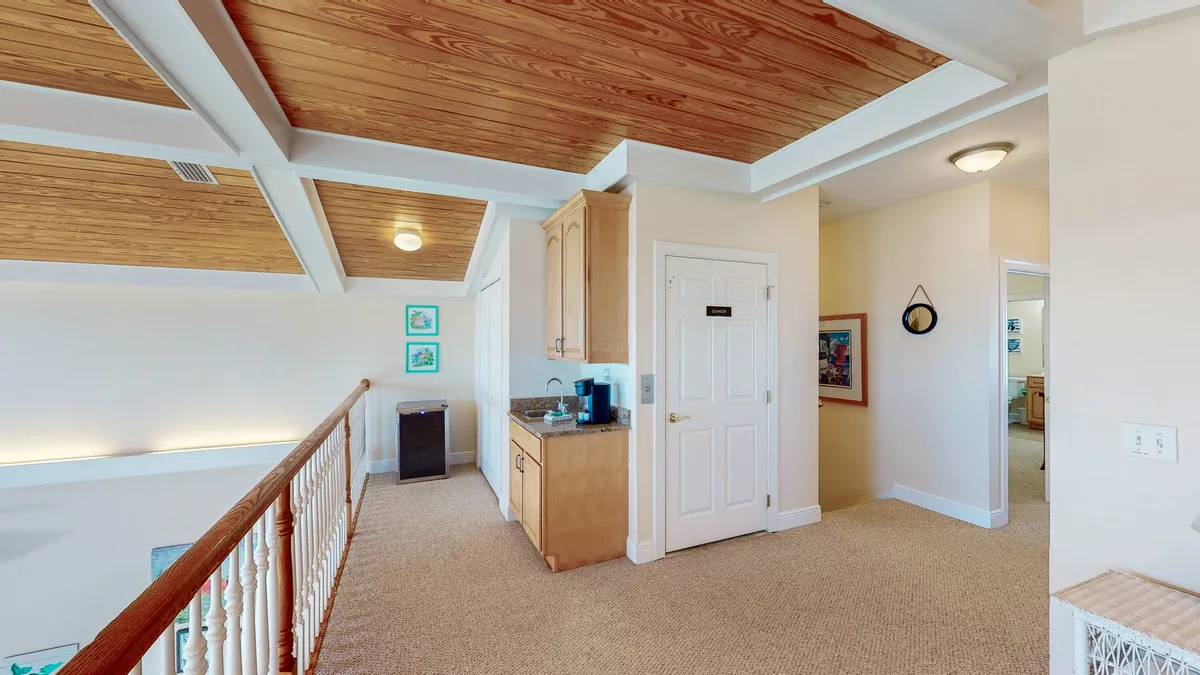 A cozy loft area featuring a small kitchenette with a coffee maker, a white door labeled 'Laundry,' and a railing overlooking the lower floor. The wooden ceiling with white beams adds warmth, making it an inviting nook for socializing or relaxing above the main living space.