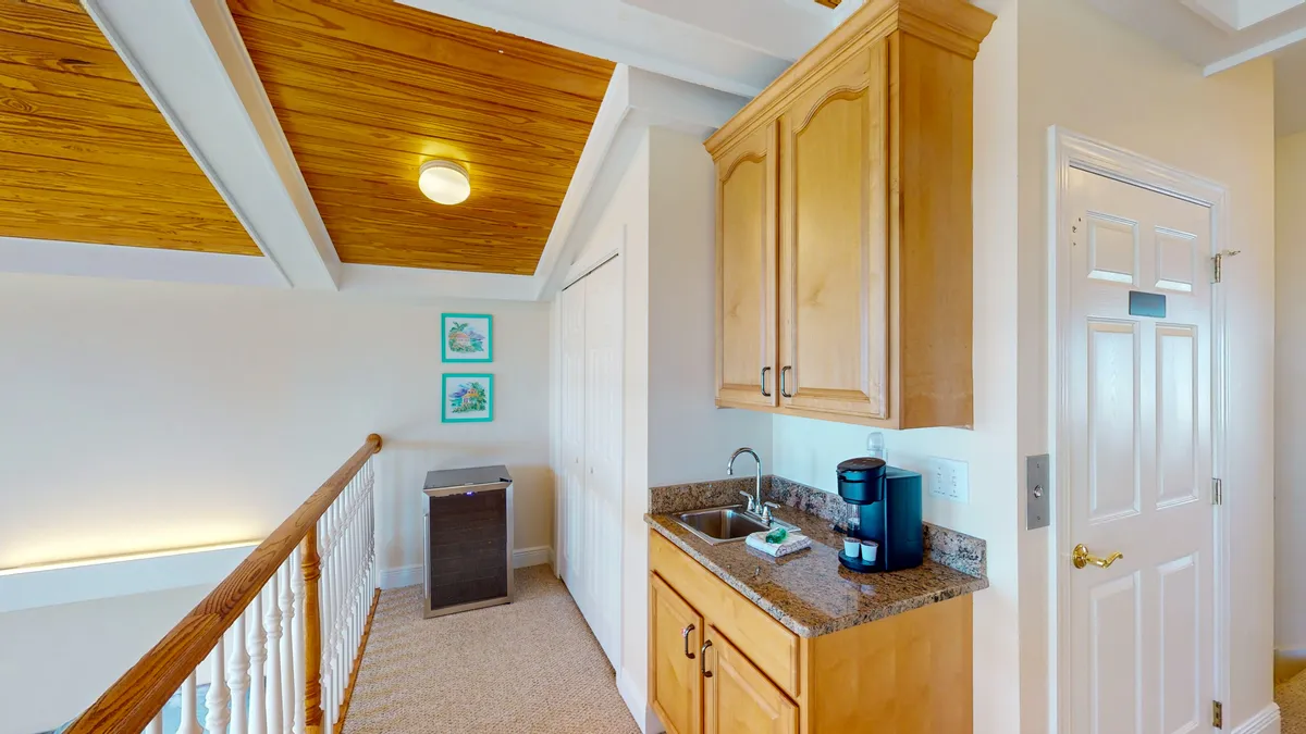 A compact kitchenette with a granite countertop, coffee maker, small black refrigerator, wood-paneled ceiling, and a white stair railing. This loft lounge wet bar area provides a relaxed spot for preparing drinks or snacks, well-suited for family hangouts or casual evenings.