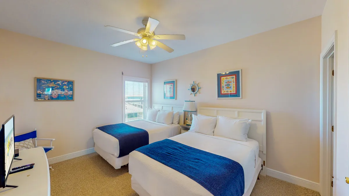 A guest bedroom with two queen beds dressed in white linens and blue throws, a ceiling fan with lights, nautical-themed wall art, and a flat-screen TV on a dresser. The space exudes calm and restful vibes with a touch of coastal charm.