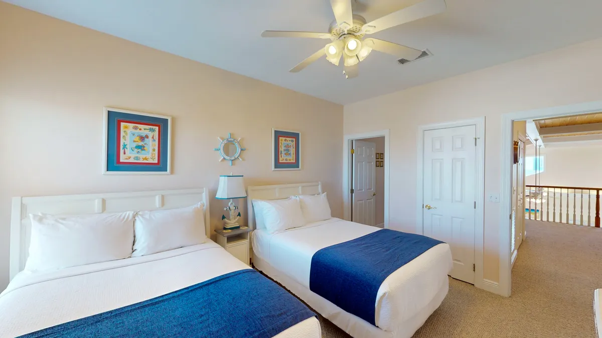 A peaceful guest bedroom with two double beds dressed in white linens and blue runners, nautical-themed wall art, a ceiling fan, and an open doorway leading to a bright loft area. The overall mood is airy and relaxed, encouraging restful sleep and easy transitions to communal spaces.