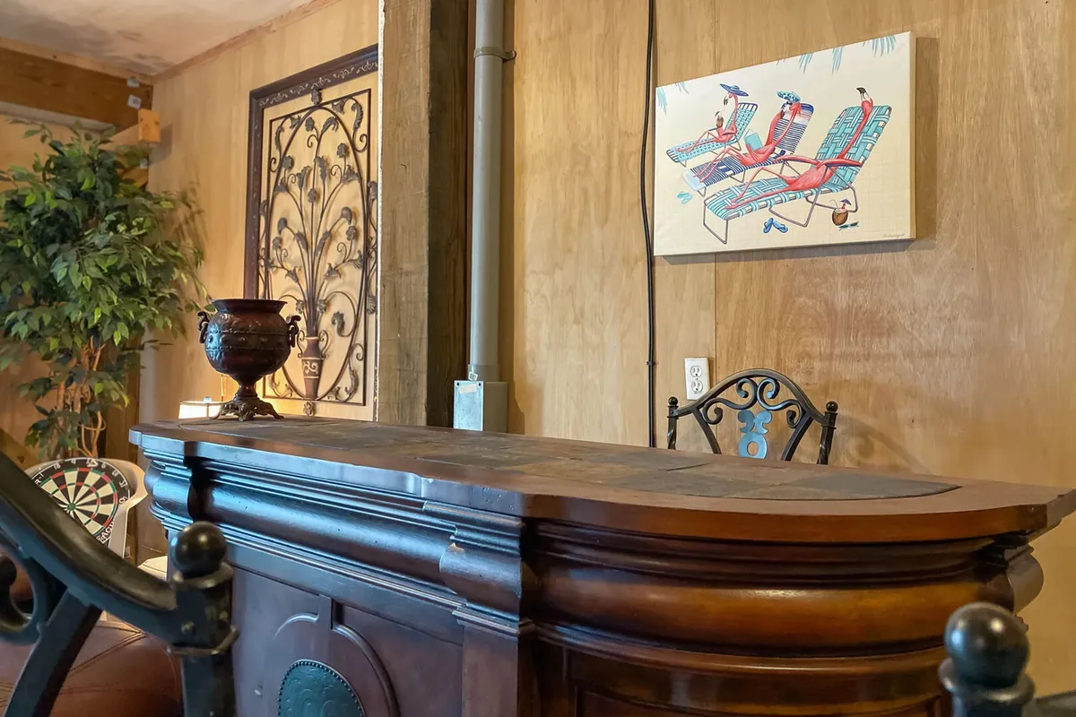 A cozy entertainment space featuring a large wooden bar with a curved design, decorative wrought iron wall art, a colorful beach-themed painting, and a dartboard partially visible behind a plant. The scene is set for socializing, playing darts, and enjoying the inviting coastal atmosphere.