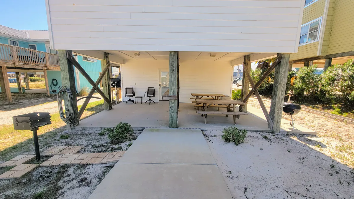 A spacious, covered patio area beneath the house features two black chairs and a small table, a classic wooden picnic table, and a barbecue grill. The patio is shaded by the home above, creating a comfortable outdoor living and dining area. The setup is casual and welcoming, ideal for barbecues, family meals, or simply relaxing in the coastal air.
