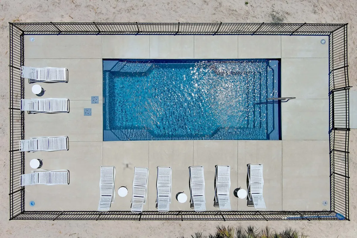 This exterior image features a sparkling rectangular private pool surrounded by white loungers and small side tables, all enclosed by a black safety fence. The area is bathed in coastal sunlight, providing a secure and inviting environment for relaxation and recreation.