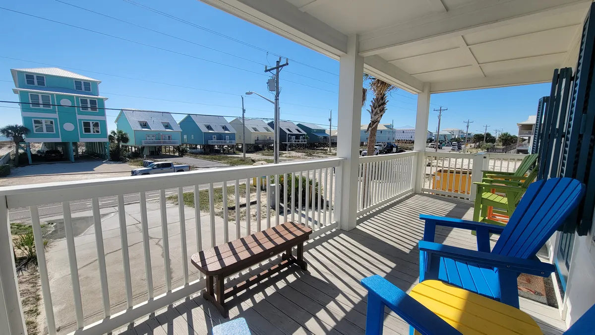 A balcony lined with colorful Adirondack chairs overlooks a quiet street of coastal homes. White railings cast patterned shadows on the wooden deck, while a palm tree and the distant neighborhood enhance the breezy, serene vibe. The balcony is perfect for morning coffee or evening sunset watching, offering a tranquil retreat with views and fresh sea air.