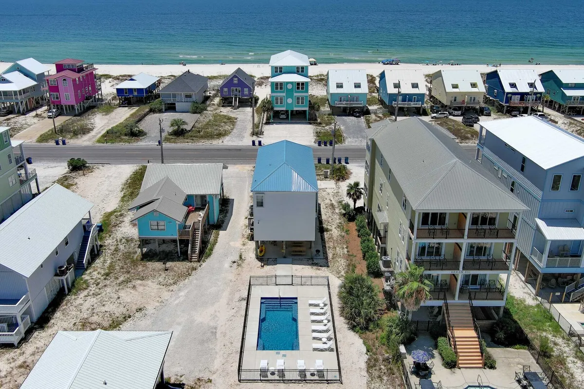 A private pool with white lounge chairs sits in the foreground, backed by stilted, colorful coastal homes and a pristine stretch of white sand beach. The calm blue Gulf waters glisten beyond, evoking a classic Gulf Shores setting. The pool area is inviting and well-maintained, ideal for swimming, sunbathing, or gathering with friends and family. The proximity of the pool to both the home and the beach is immediately evident.
