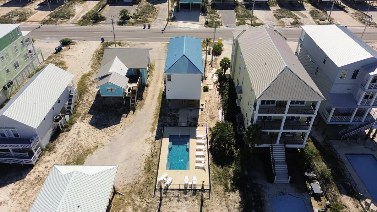 This exterior shot features the private swimming pool surrounded by lounge chairs, with the distinctive coastal home (blue roof) and neighboring beach houses visible. The sandy driveway and open yard evoke a laid-back vacation atmosphere, and the pool is showcased as a central feature for outdoor fun.