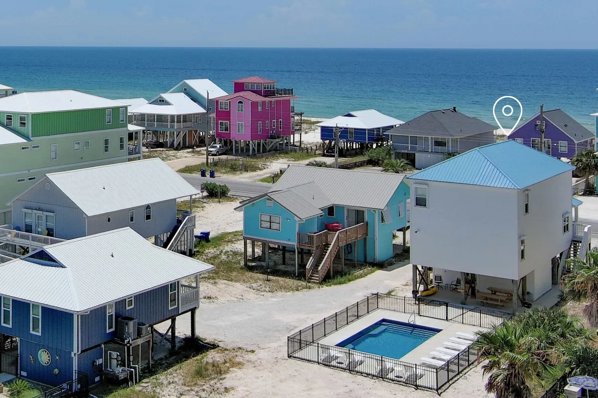 A cheerful outdoor scene features a private pool enclosed by a black metal fence, with colorful raised beach houses and the Gulf ocean horizon in the background. Staircases lead up to inviting homes, and the pool area is ideal for relaxing after time on the sand.