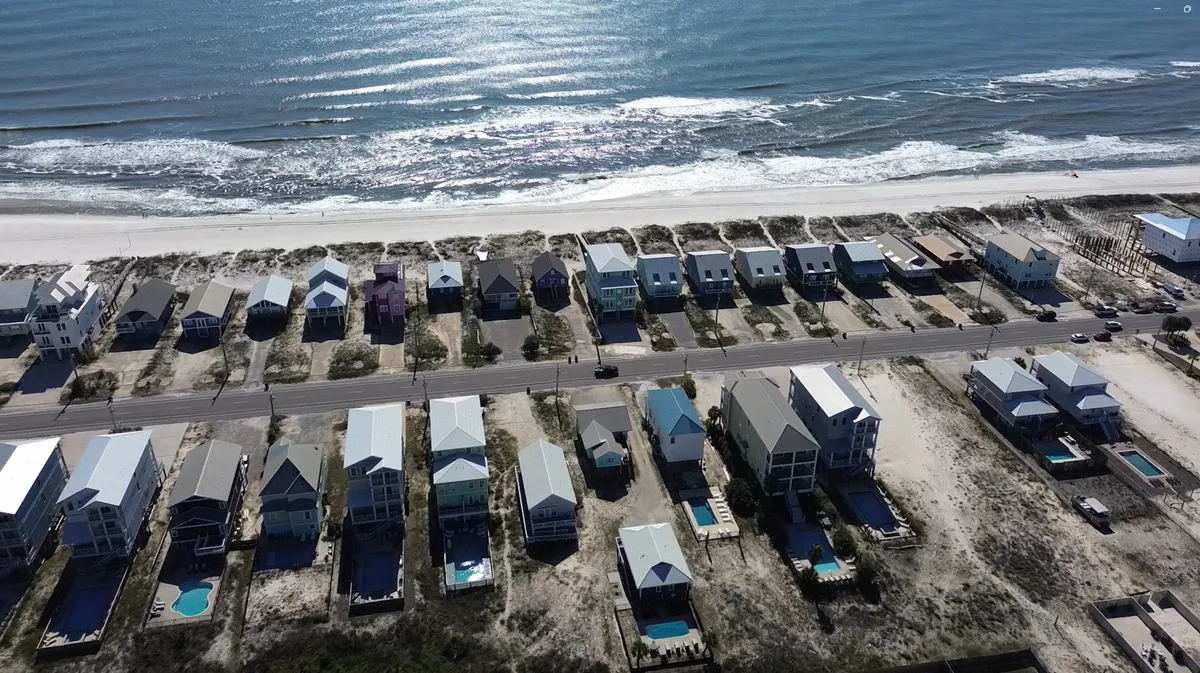 A sweeping exterior view of beachfront homes with private pools, white sandy beach, calm ocean waves, and a coastal road separating the homes from the beach. The scene is tranquil, offering a panoramic sense of Gulf Shores’ beauty and proximity to the water.
